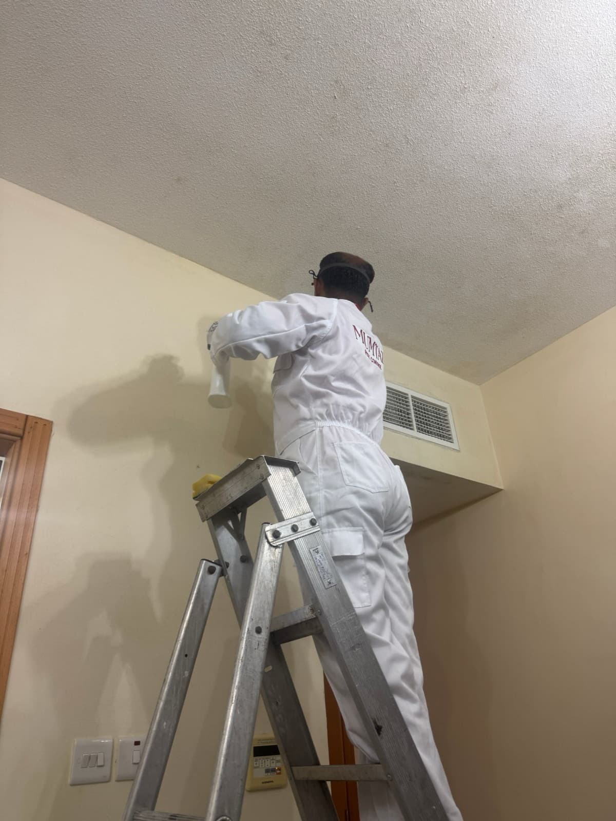 Technician in white coveralls on a stepladder treating the ceiling-to-wall junction beside an AC grille